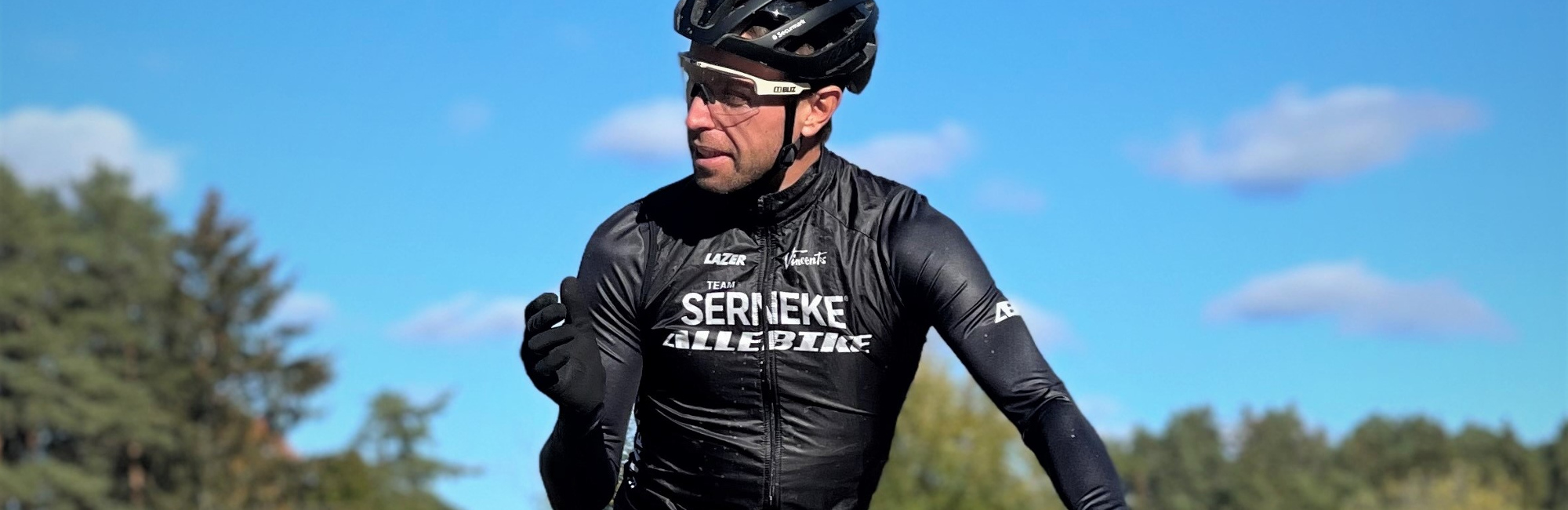 MTB-cyklisten Emil Lindgren från Team Serneke-Allebike i cykelkläder, cykelglasögon och Lazer cykelhjälm på en vit cykel med blå himmel som bakgrund.