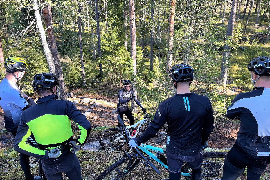 Emil Lindgren med cykelkläder, svart Lazer hjälm, vit MTB-cykel med Shimano XTR och PRO-komponenter som undervisar fyra mountainbikecyklister i skogen. 