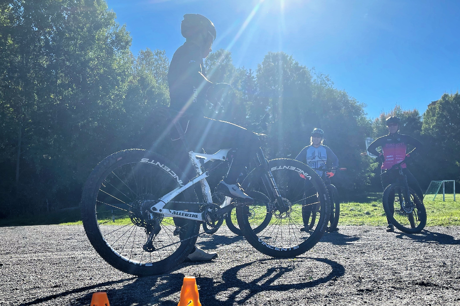 Emil Lindgren med cykelkläder, svart Lazer hjälm, vit MTB-cykel med Shimano XTR och PRO-komponenter samt orangefärgade koner på marken i motljus framför deltagarna på MTB-clinic. 