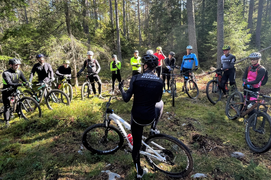 Emil Lindgren med svarta cykelkläder och Lazer hjälm, samt vit MTB-cykel med Shimano XTR och PRO-komponenter, står med ryggen mot kameran framför sin grupp med deltagare på MTB-clinic i skogen.
